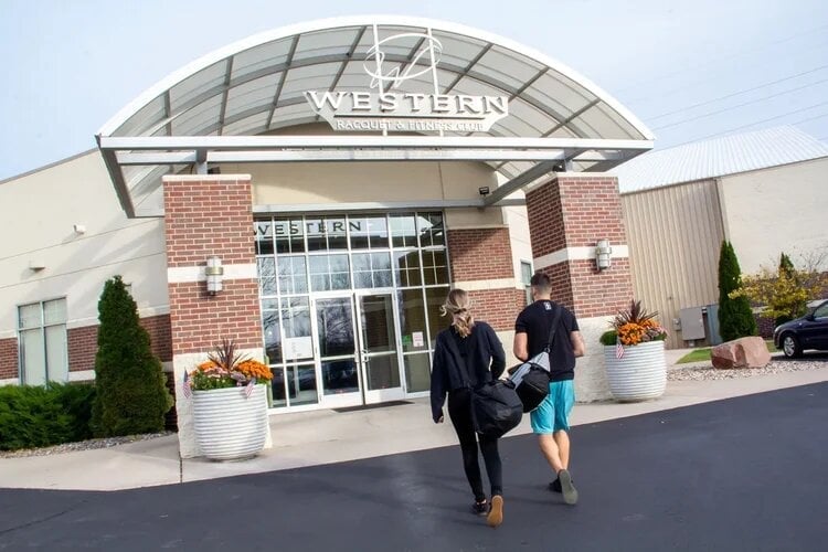 Two people walking toward a Western-branded entrance building with a curved metal roof, brick facade, and planters flanking the glass doors.